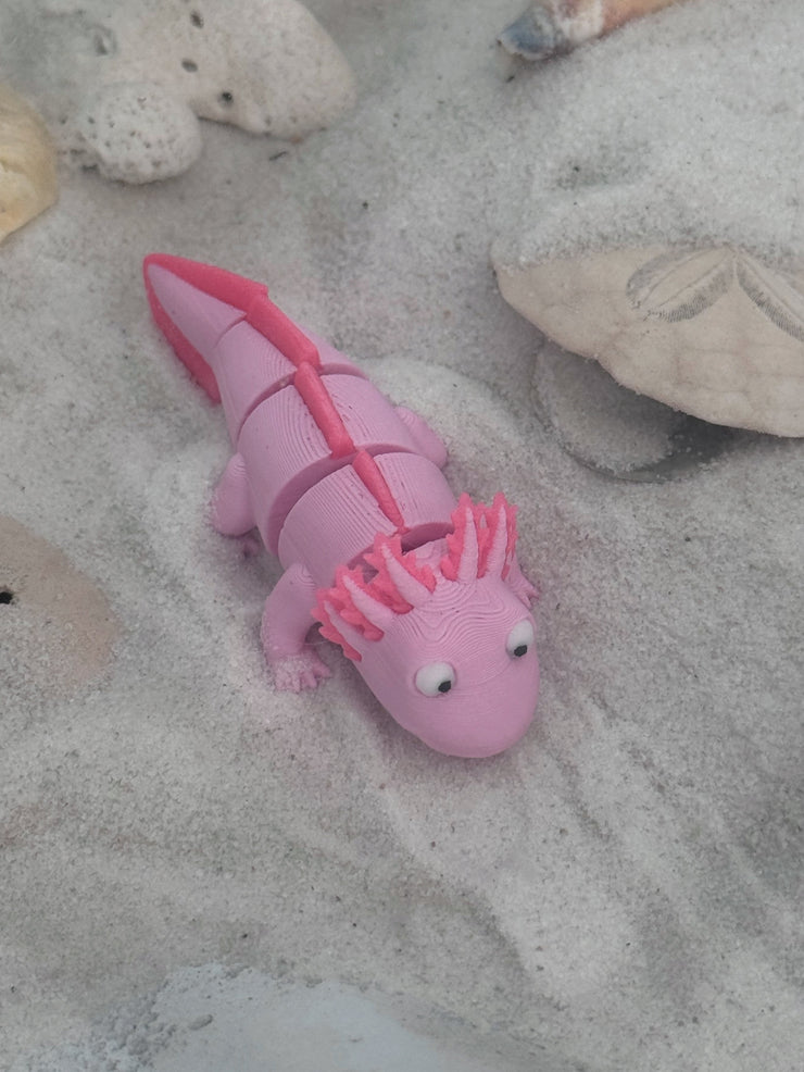 Pink axolotl toy on sandy surface with shells and sandcastles in the background